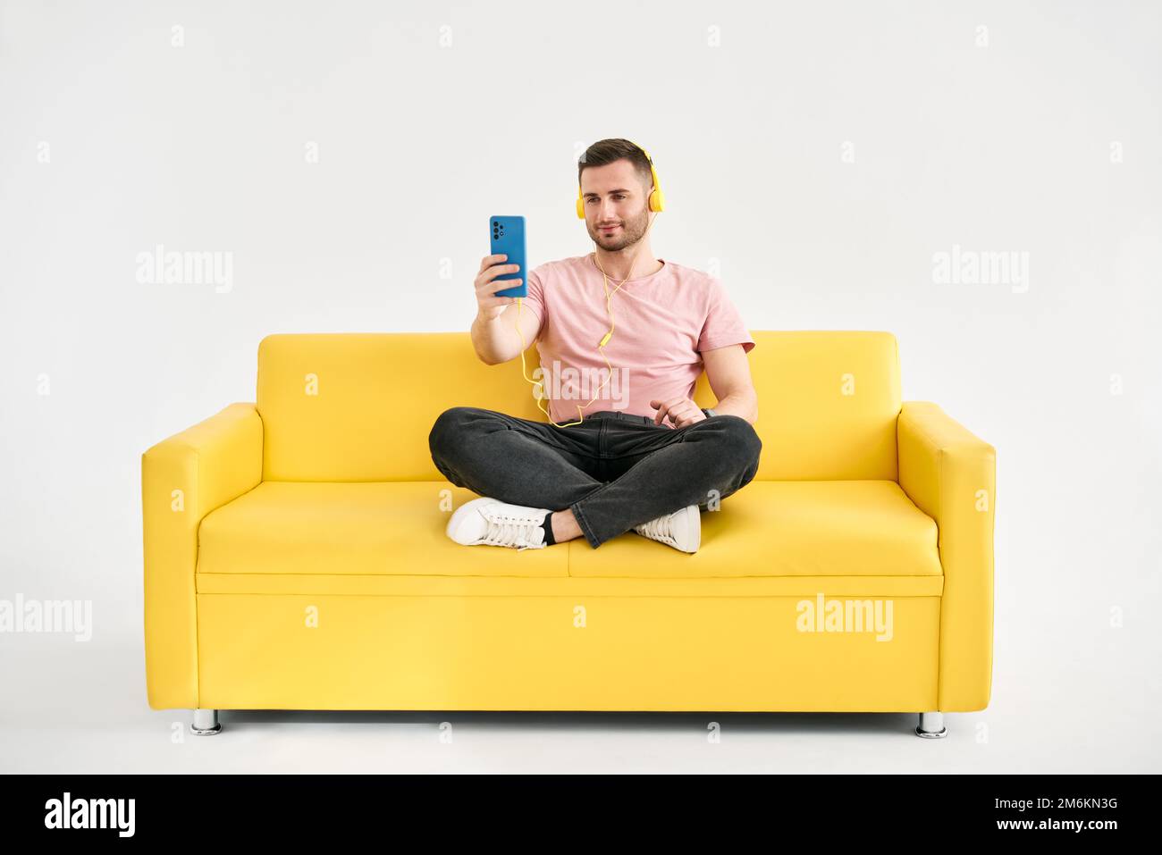 Décontracté jeune homme porter un casque écouter de la musique sur son téléphone portable tout en étant assis sur un canapé jaune Banque D'Images