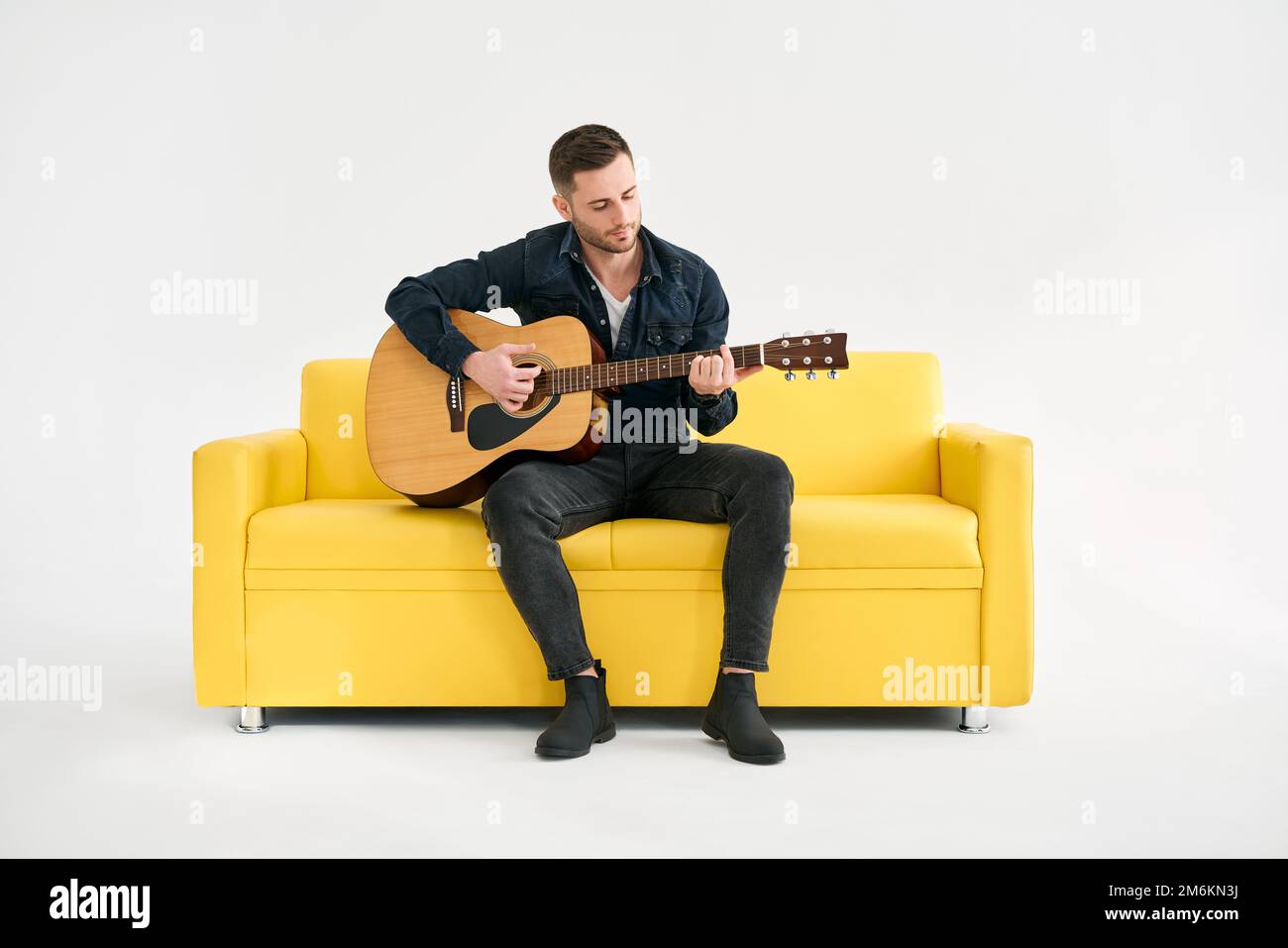 Beau jeune homme jouant de la guitare acoustique whlile assis sur un canapé jaune sur fond blanc Banque D'Images