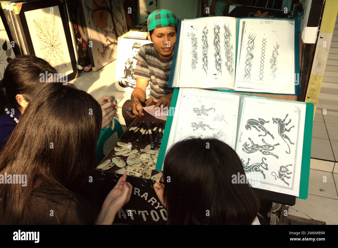 Un shopper attend alors qu'un groupe de femmes touristes discutent d'options tout en se croquant devant un grand livre de présentation de tatouage dans un magasin d'art côté rue à Tanah Lot, Tabanan, Bali, Indonésie. Banque D'Images