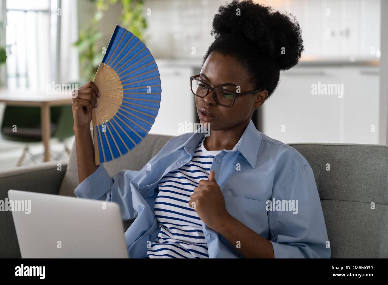 Femme en surchauffe épuisée souffrant de coup de chaleur agitant le ventilateur et regardant l'ordinateur portable se trouve sur le canapé Banque D'Images