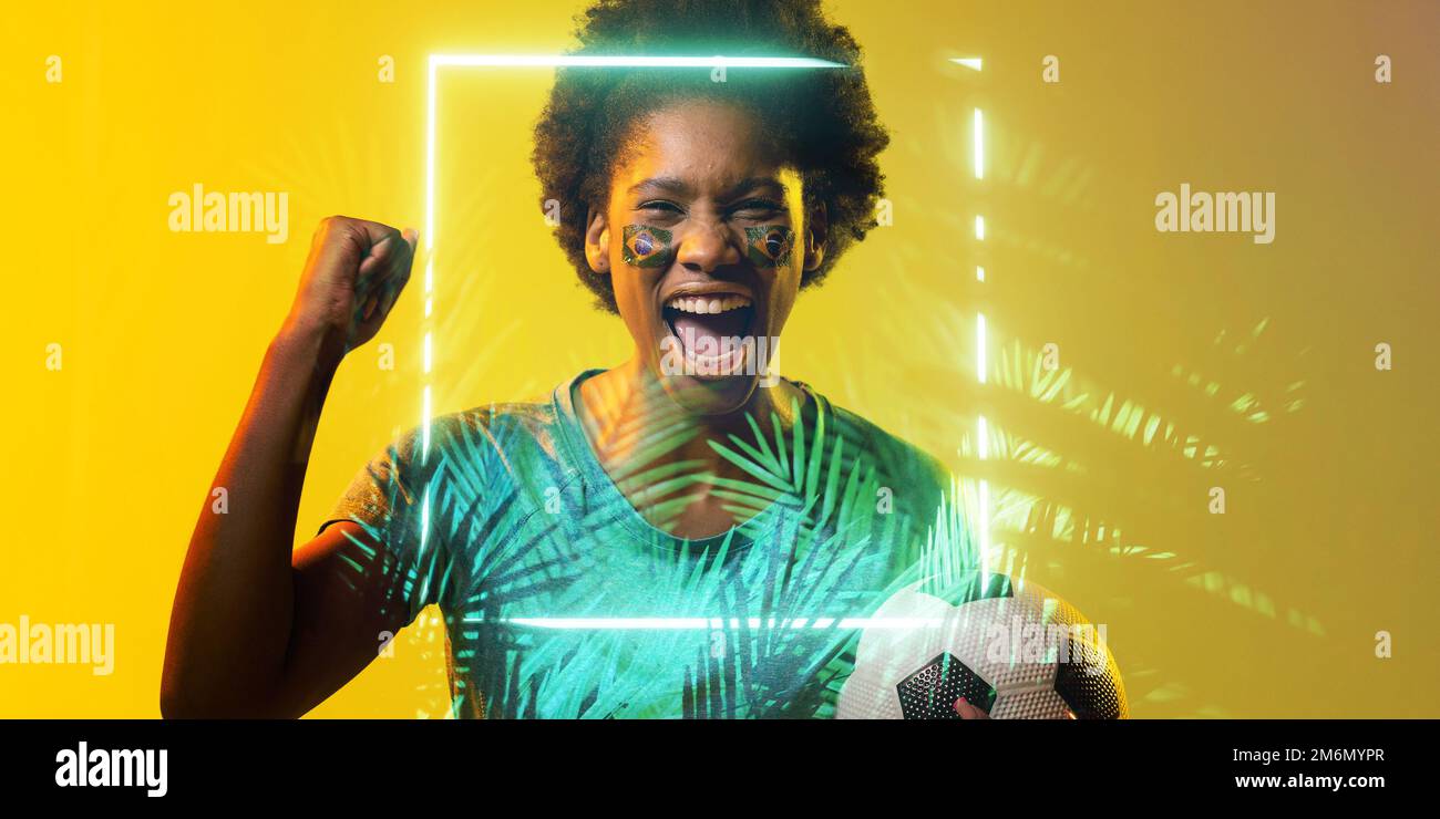 Fan afro-américain avec le visage du drapeau brésilien peint criant par le rectangle et les plantes Banque D'Images