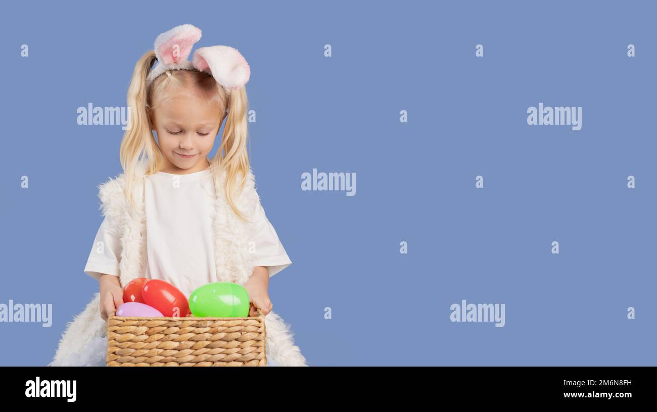Belle petite fille mignonne dans les oreilles de lapin. L'enfant tient dans les mains et regarde dans le panier de Pâques avec des œufs sur fond bleu. Copier l'espace. Banque D'Images