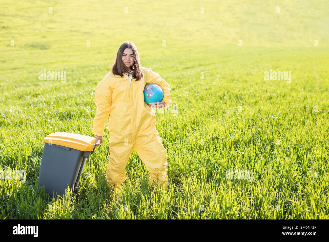 Une femme dans un costume de protection jaune se tient au milieu d'un champ vert tenant un globe, et avec son autre main porte une poubelle. Banque D'Images