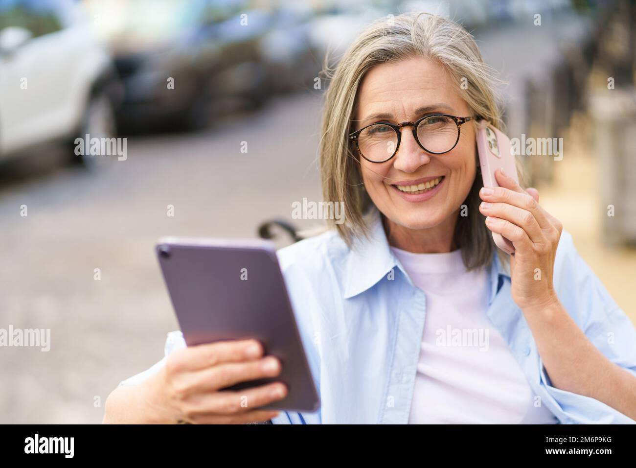 Lire les mises à jour sur la tablette numérique mature cheveux gris femme parlant au téléphone assis sur le banc dans les rues de l'ancienne Europea Banque D'Images