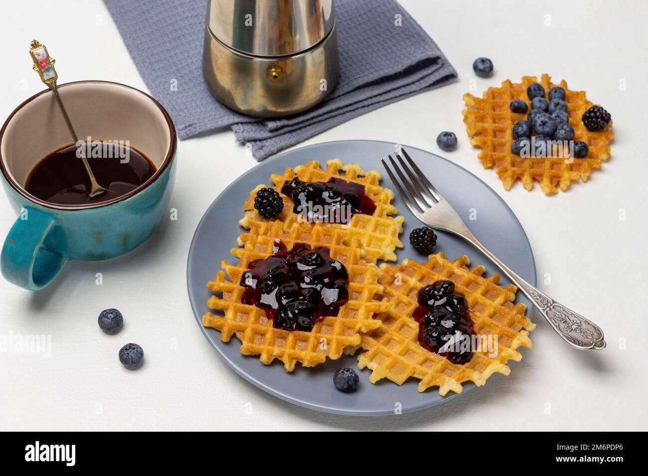 Gaufres avec confiture et fourchette sur assiette grise. Une tasse de café bleu et une cafetière sur une serviette grise. Vue de dessus. Arrière-plan blanc. Banque D'Images