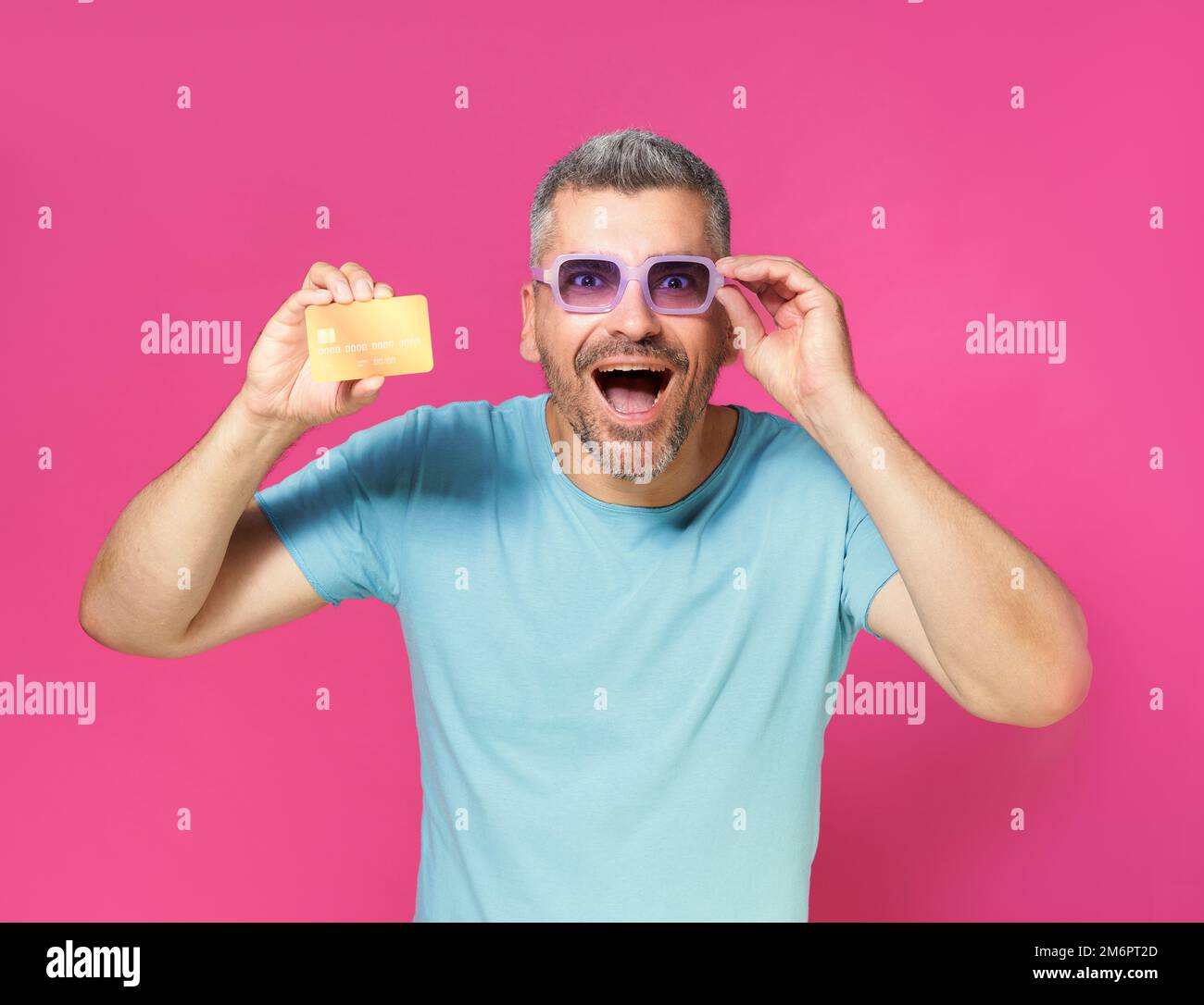 Excité, choqué beau homme d'âge moyen tiennent le débit, carte de crédit à la main fixant ses lunettes portant le t-shirt bleu isolé sur pi Banque D'Images
