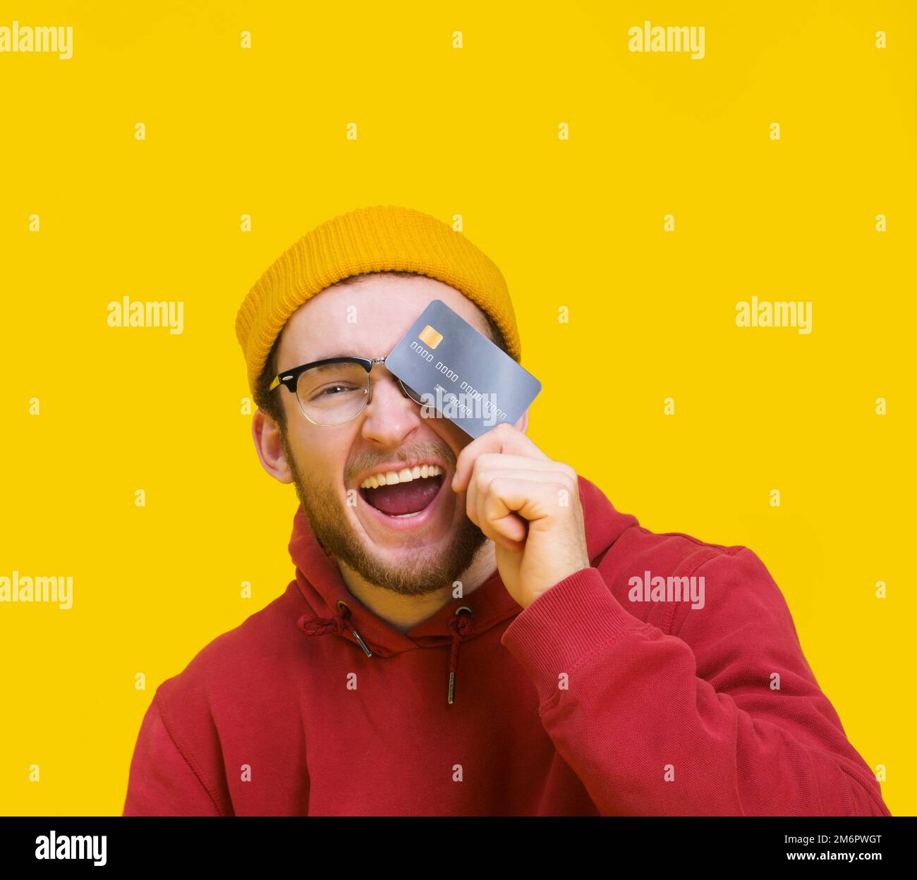 Jeune homme avec carte de crédit ou de débit dans la main souriant heureux couvrir son oeil prêt à dépenser de l'argent. Jeune homme à capuchon rouge avec mocku Banque D'Images