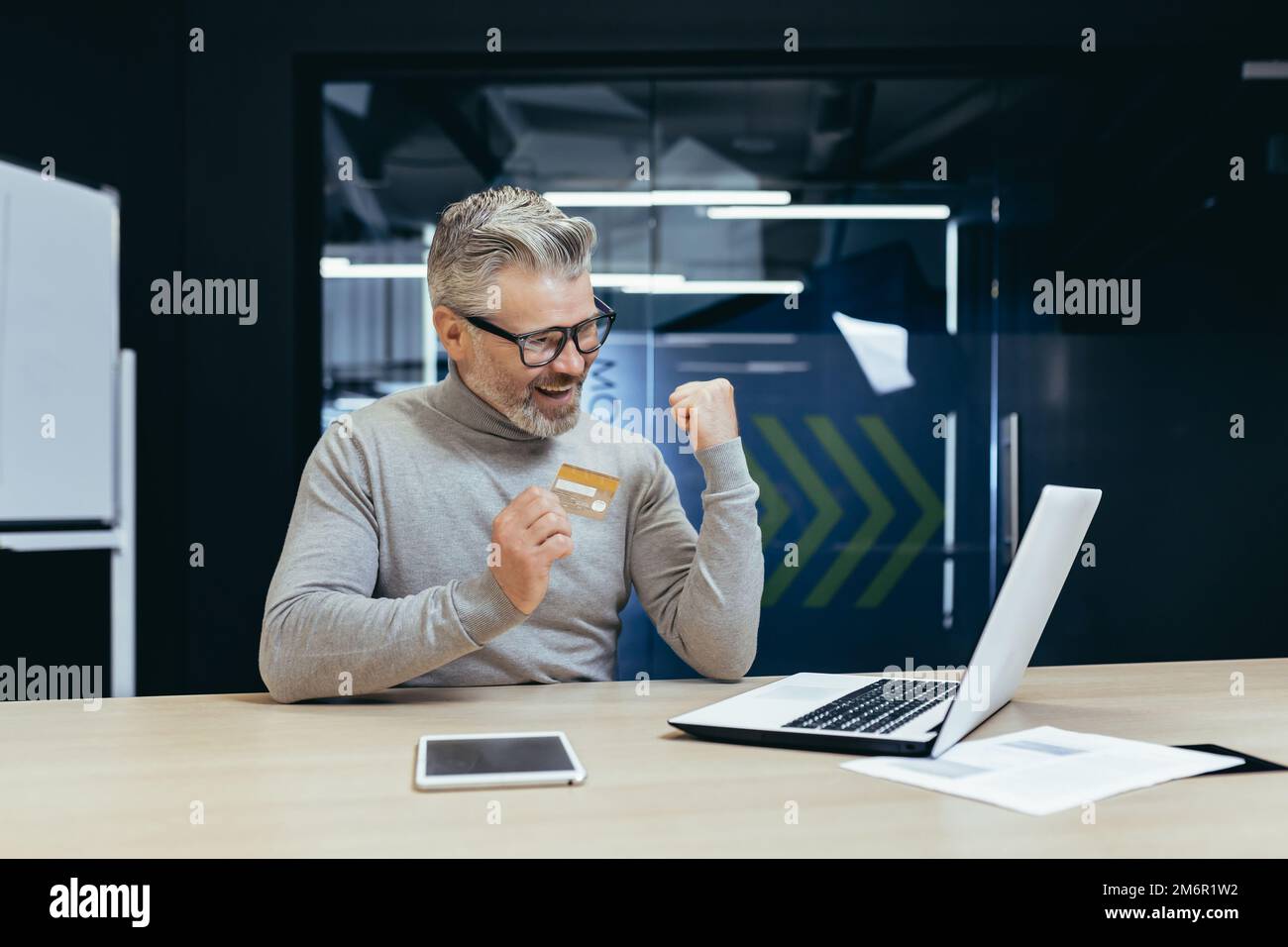 Homme d'affaires heureux et souriant au bureau faisant le transfert bancaire et l'achat en ligne dans la boutique en ligne, homme mature tenant la carte bancaire de crédit et utilisant l'ordinateur portable, heureux avec le choix. Banque D'Images