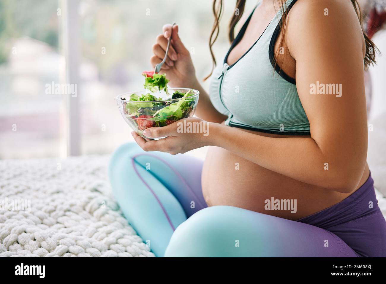 Gros plan portrait d'une femme enceinte en vêtements de sport mangeant une salade fraîche assise au lit Banque D'Images