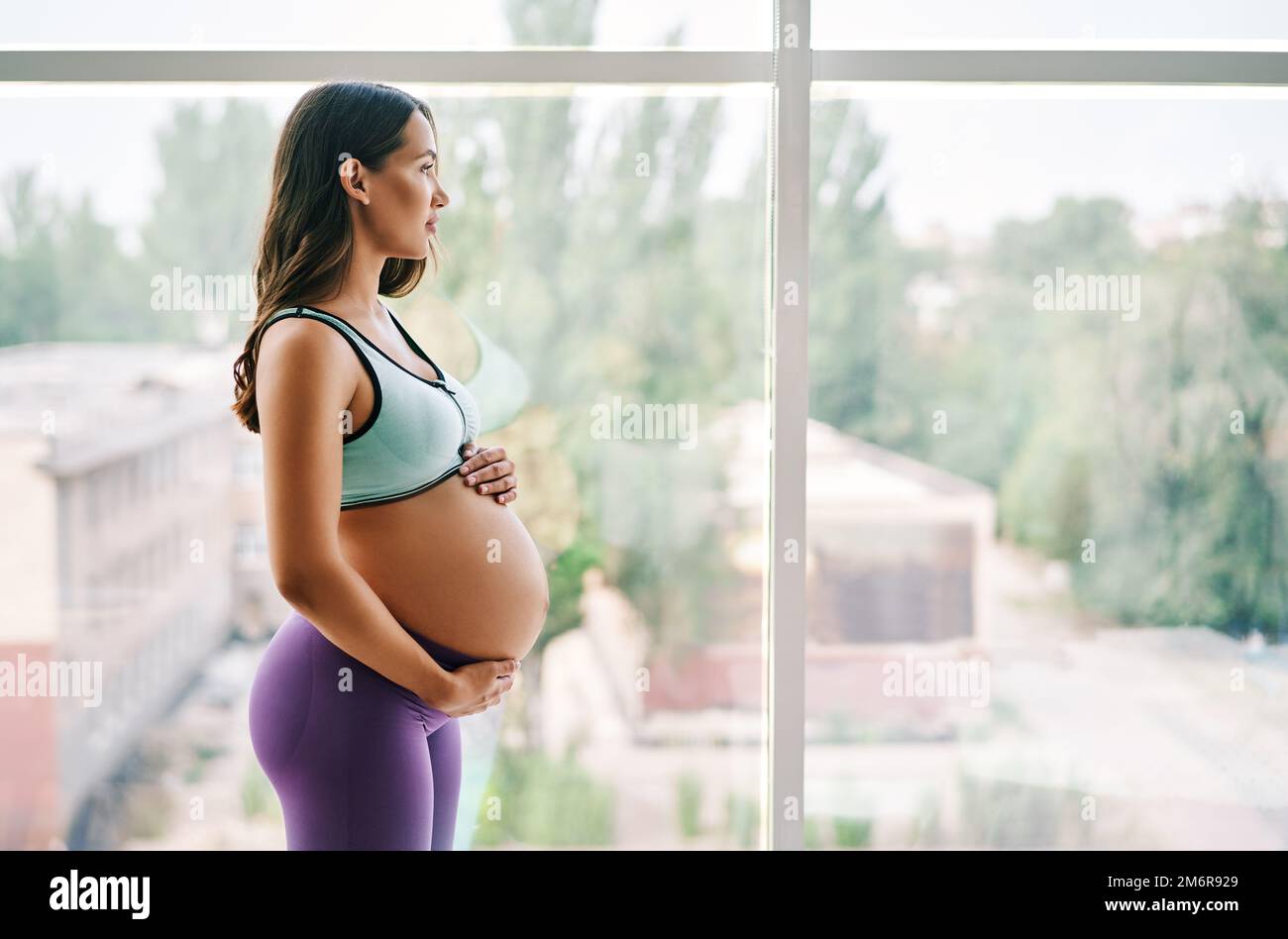 Portrait d'une jeune femme enceinte dans des vêtements de sport debout près de la fenêtre tenant les mains sur son ventre avec l'espace de copie Banque D'Images