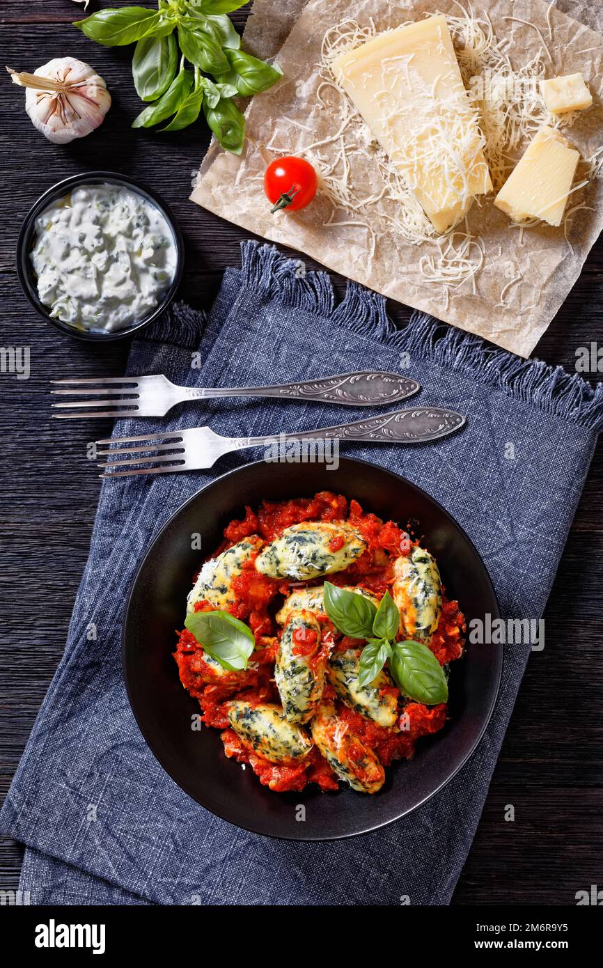 Boulettes de ricotta aux épinards italiens sauce tomate aux herbes et parmesan râpé dans un bol noir sur une table en bois sombre avec ingrédients Banque D'Images