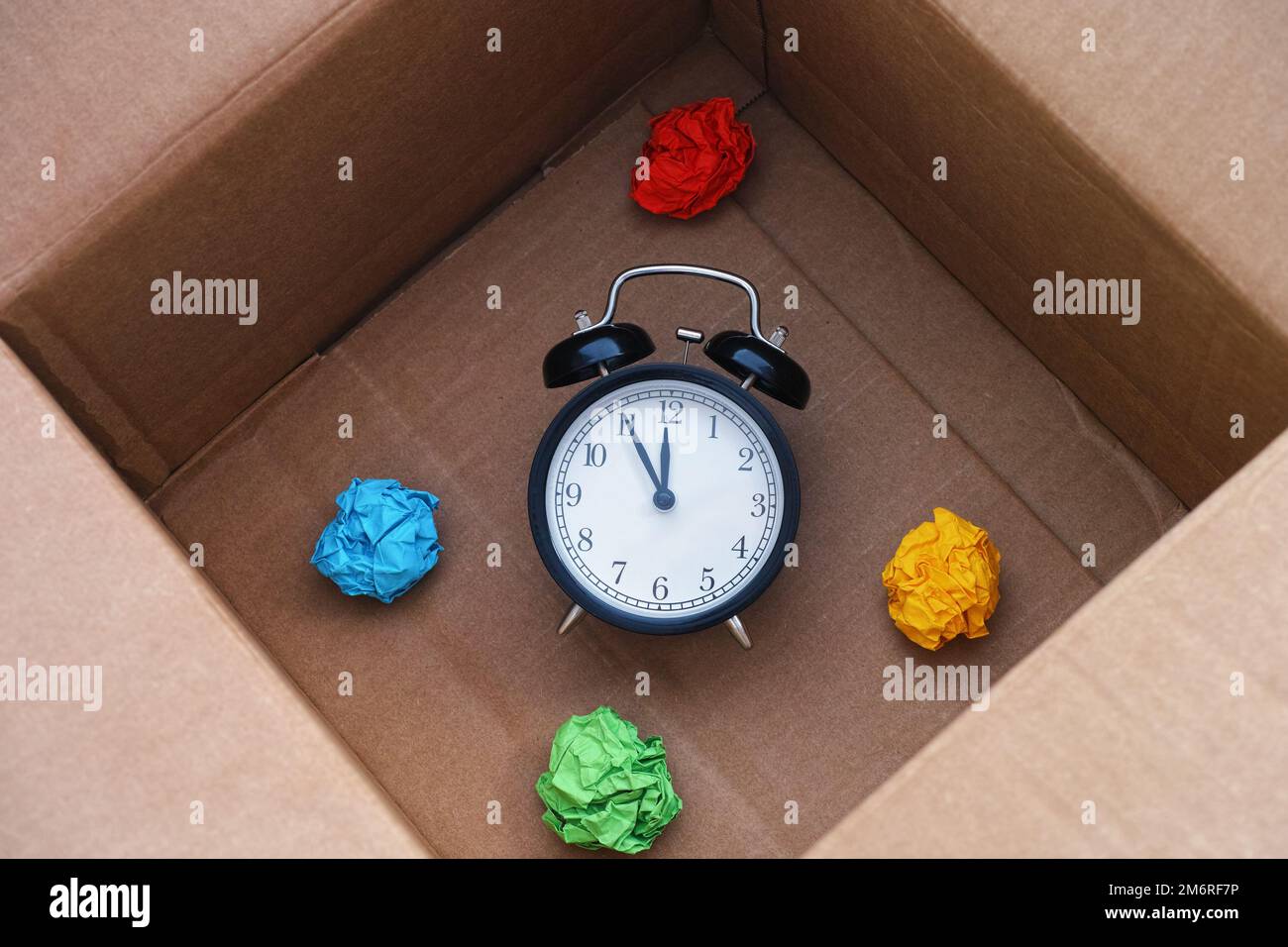 Un réveil placé dans une boîte en carton à côté de boules de papier froissé de couleur. Concept de temps écoulé ou d'échéance. Banque D'Images