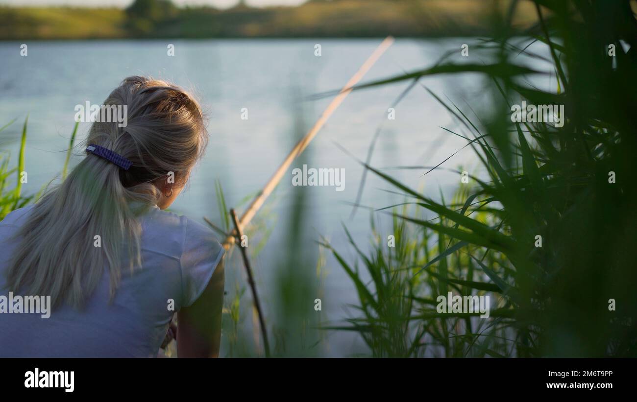 Une jeune fille qui pêche, assise sur la rive avec une canne à pêche et attendant une prise. Une jeune fille pêche depuis un quai dans un lac par une journée ensoleillée Banque D'Images