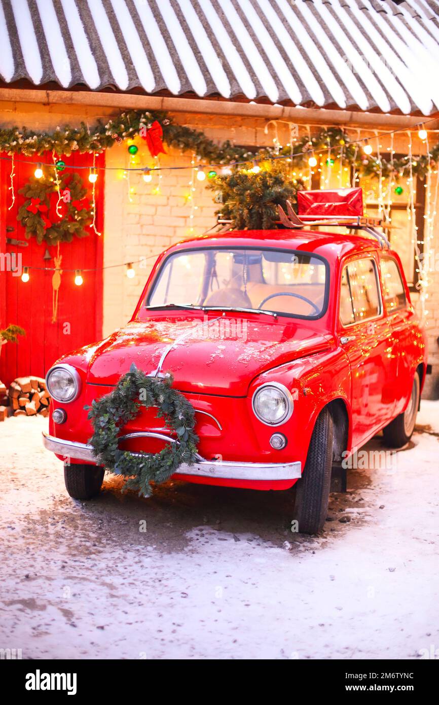 Voiture rétro rouge dans la cour couverte de neige de la maison décorée avec des branches de pin et des lumières de Noël. Banque D'Images
