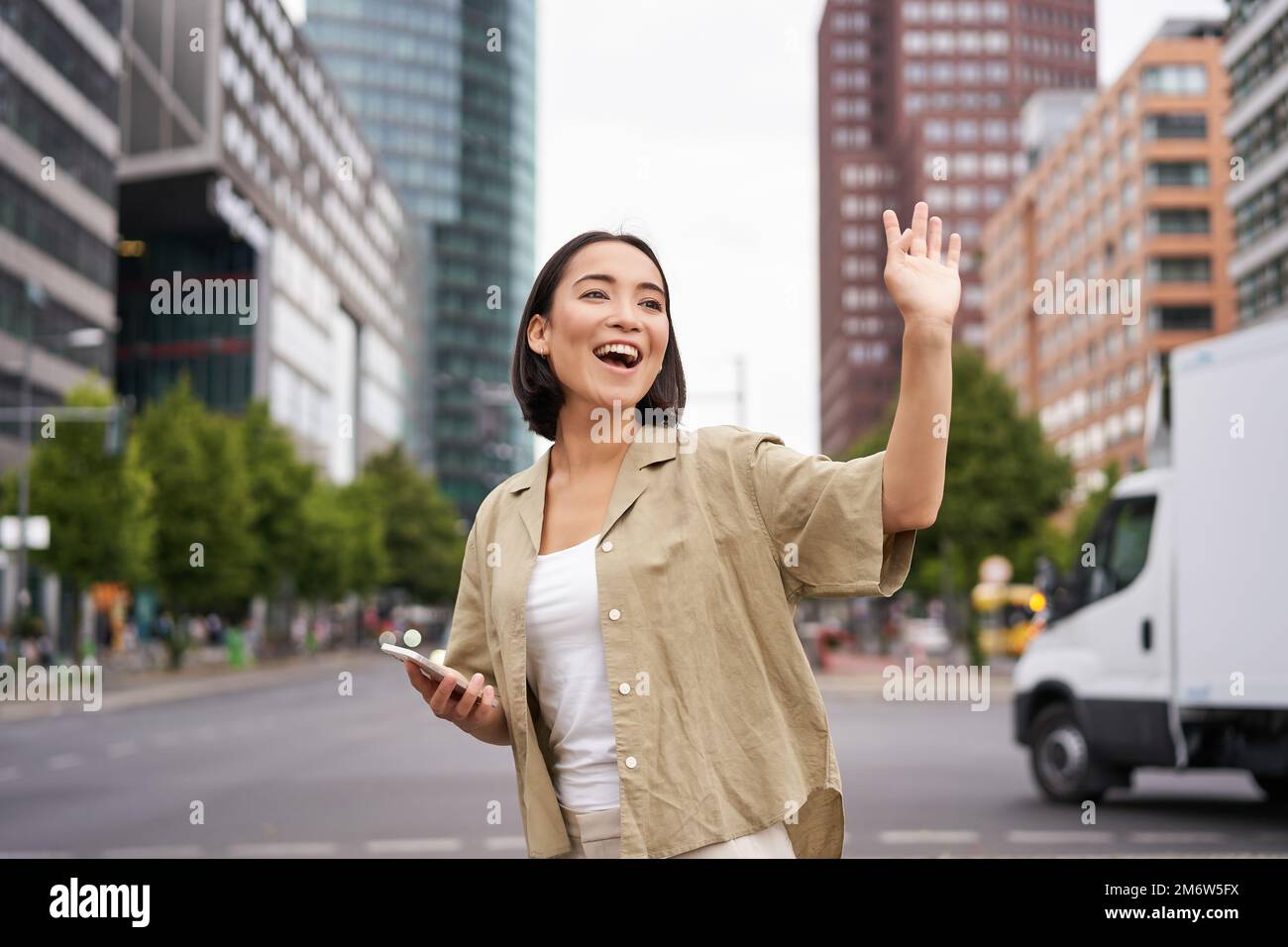 Bonne fille asiatique passant par un ami et leur faisant signe dans la rue, en disant bonjour tout en marchant dans la ville, en tenant un smartphone Banque D'Images