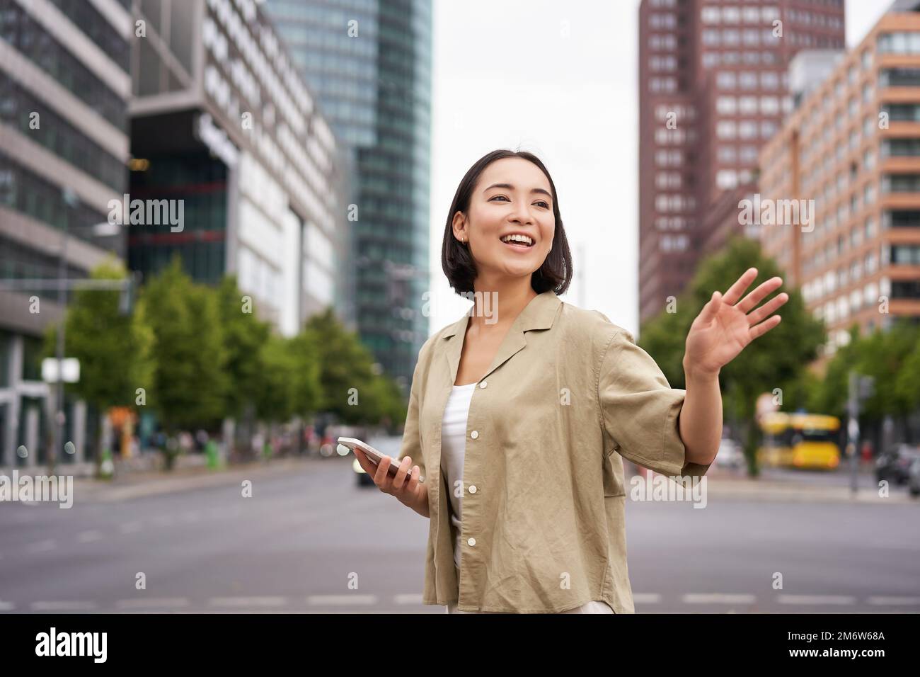 Bonne fille asiatique passant par un ami et leur faisant signe dans la rue, en disant bonjour tout en marchant dans la ville, en tenant un smartphone Banque D'Images