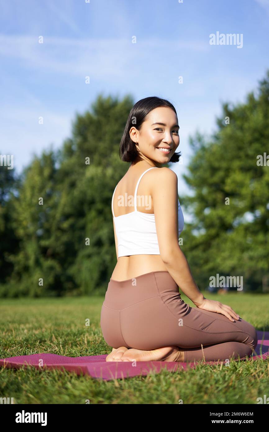 Portrait d'une femme asiatique assise et méditant dans le parc, faisant du yoga en plein air, des exercices de remise en forme, souriant à l'appareil photo Banque D'Images