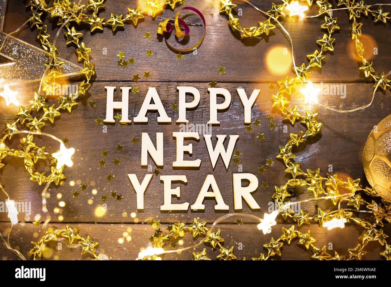 Joyeux nouvel an, lettres en bois sur un fond festif avec des paillettes, des étoiles, des lumières de guirlandes. Salutations, carte postale. Banque D'Images