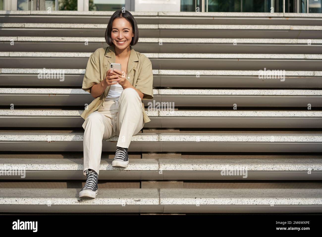 Portrait d'une fille asiatique souriante assis sur un escalier avec son smartphone, en naviguant sur Internet sur un téléphone mobile, se reposant à l'extérieur dans la ville c Banque D'Images