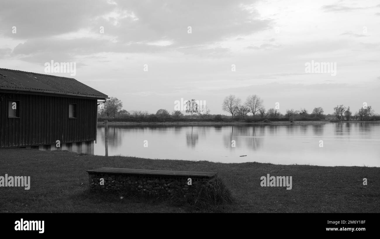 Banc rempli de pierres derrière les grilles sur la petite rive au lac de constance après le coucher du soleil avec hutte en bois à gauche en noir et blanc Banque D'Images