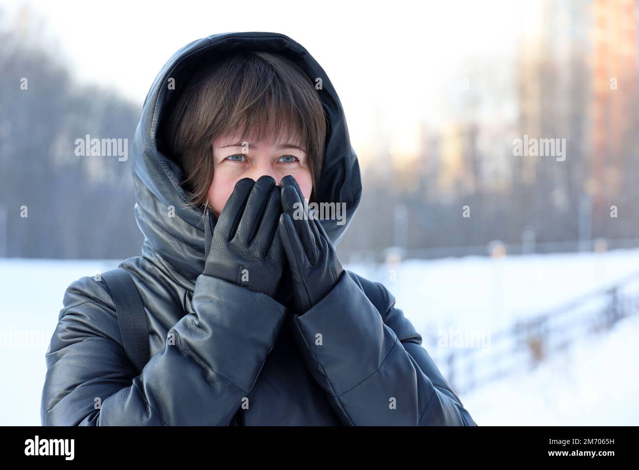 Temps de gel en hiver, femme en cuir manteau avec capuche en fourrure marchant dans une rue pendant la neige et couvrant son visage par les mains dans des gants Banque D'Images
