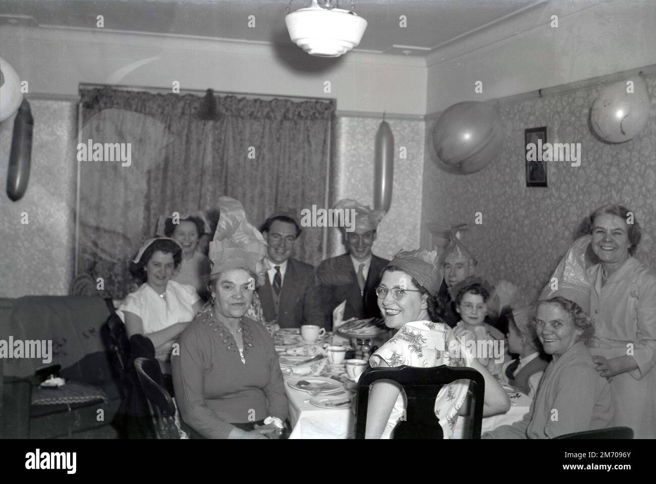 1950s, historique, fête de famille, grands sourires en famille, la plupart portant des chapeaux de fête, s'asseoir dans une salle de devant autour d'une table prenant un repas ensemble, ballons sur les wallls, Angleterre, Royaume-Uni. Banque D'Images