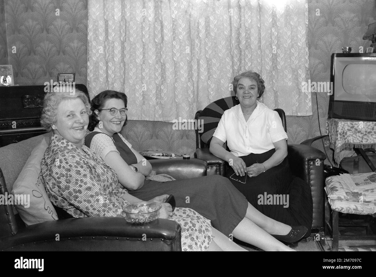 1950s, historique, trois dames assises dans une salle de réception, un ensemble de télévision de l'époque au coin, Angleterre, Royaume-Uni, une télévision EKCO. Fondée en 1924, à Southend-on-Sea, EKCO est une société d'électronique britannique, nommée d'après son fondateur Eric Kirkham Cole. E. K Cole Ltd, a commencé à fabriquer des postes de radio, puis, pendant la Seconde Guerre mondiale, a développé un système radar d'avertissement. La production de téléviseurs a commencé au début de 50s et, en 1957, ils avaient fabriqué un million de téléviseurs. EKCO a fusionné avec une autre société britannique d'électronique, la société PYE de cambridge en 1960 pour former British Electronics Industries Ltd Banque D'Images 1950s, historique, trois dames assises dans une salle de réception, un ensemble de télévision de l'époque au coin, Angleterre, Royaume-Uni, une télévision EKCO. Fondée en 1924, à Southend-on-Sea, EKCO est une société d'électronique britannique, nommée d'après son fondateur Eric Kirkham Cole. E. K Cole Ltd, a commencé à fabriquer des postes de radio, puis, pendant la Seconde Guerre mondiale, a développé un système radar d'avertissement. La production de téléviseurs a commencé au début de 50s et, en 1957, ils avaient fabriqué un million de téléviseurs. EKCO a fusionné avec une autre société britannique d'électronique, la société PYE de cambridge en 1960 pour former British Electronics Industries Ltd Banque D'Images