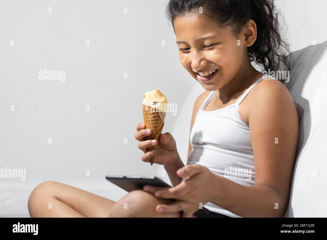 une petite fille brune heureuse et souriante mangeant une glace tout en jouant sur sa tablette ou son téléphone portable, assise sur le canapé dans son salon. Banque D'Images