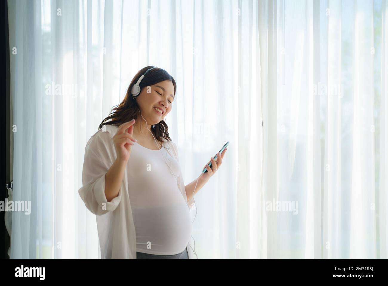 Portrait d'une femme asiatique heureuse enceinte debout dans le salon à la maison et utilisant un smartphone pour écouter de la musique dans un casque. Banque D'Images