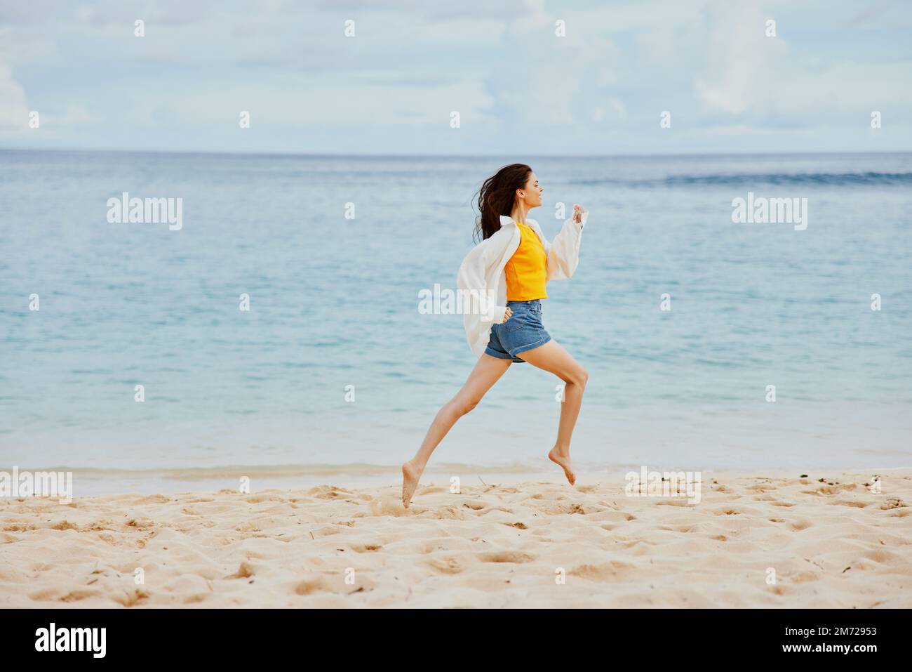 Une femme sportive court le long de la plage en vêtements d'été sur le sable dans un t-shirt jaune et un short en Jean blanc shirt volant cheveux vue sur l'océan Banque D'Images