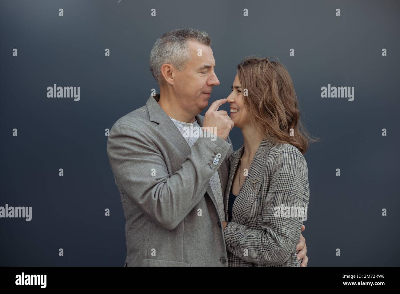 Couple romantique, homme mûr aux cheveux gris et jeune femme embrassant, souriant et touchant le nez contre le mur gris à l'extérieur Banque D'Images