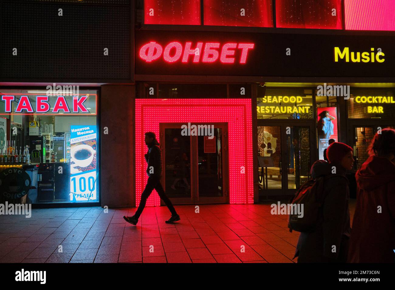 Un jeune homme qui marche le long du trottoir passe par les fenêtres des magasins illuminées la nuit. Rue Novy Arbat (New Arbat), Moscou, Russie. Banque D'Images