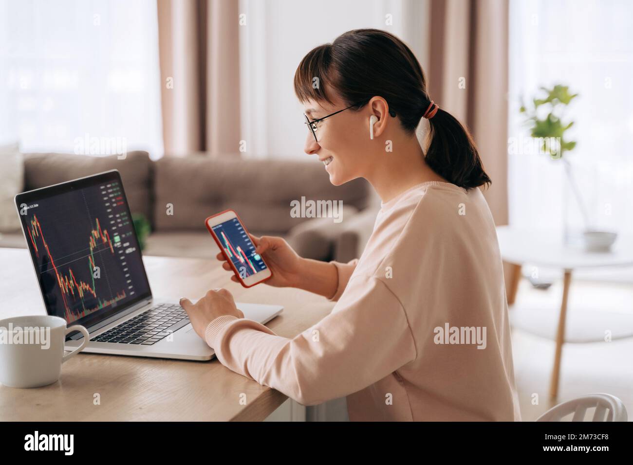 Femme d'affaires souriante négociant analyste regardant l'écran d'ordinateur portable, tenant le smartphone, portant des écouteurs. Investisseur courtier analysant des indices, négociant des données d'investissement en ligne sur le graphique de marché boursier à la maison Banque D'Images
