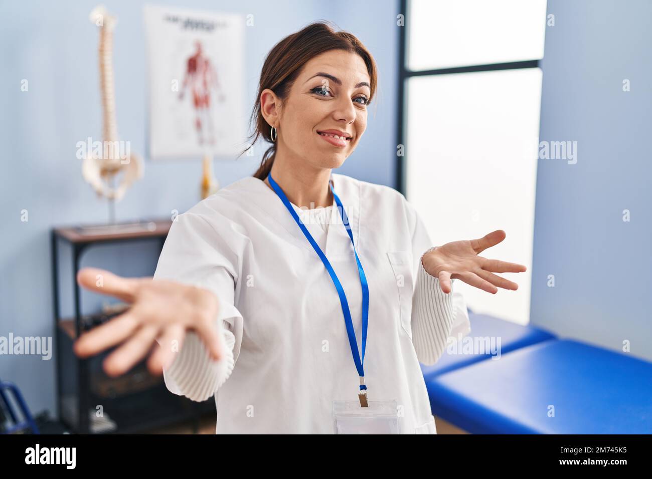 Jeune femme brunette travaillant à la clinique de rétablissement de la douleur souriant gai avec les bras ouverts comme accueil amical, positif et confiant salutations Banque D'Images