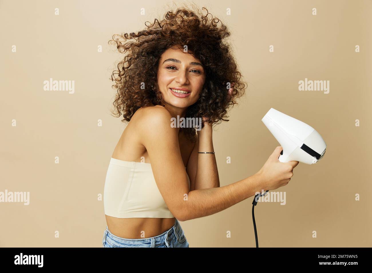 Femme sèche les cheveux freux avec sèche-cheveux, maison beauté produits coiffants cheveux, sourire sur fond beige Banque D'Images