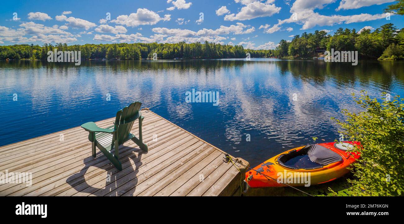 Chaise assise sur un quai en bois faisant face à un lac calme avec un canoë rouge en été Banque D'Images
