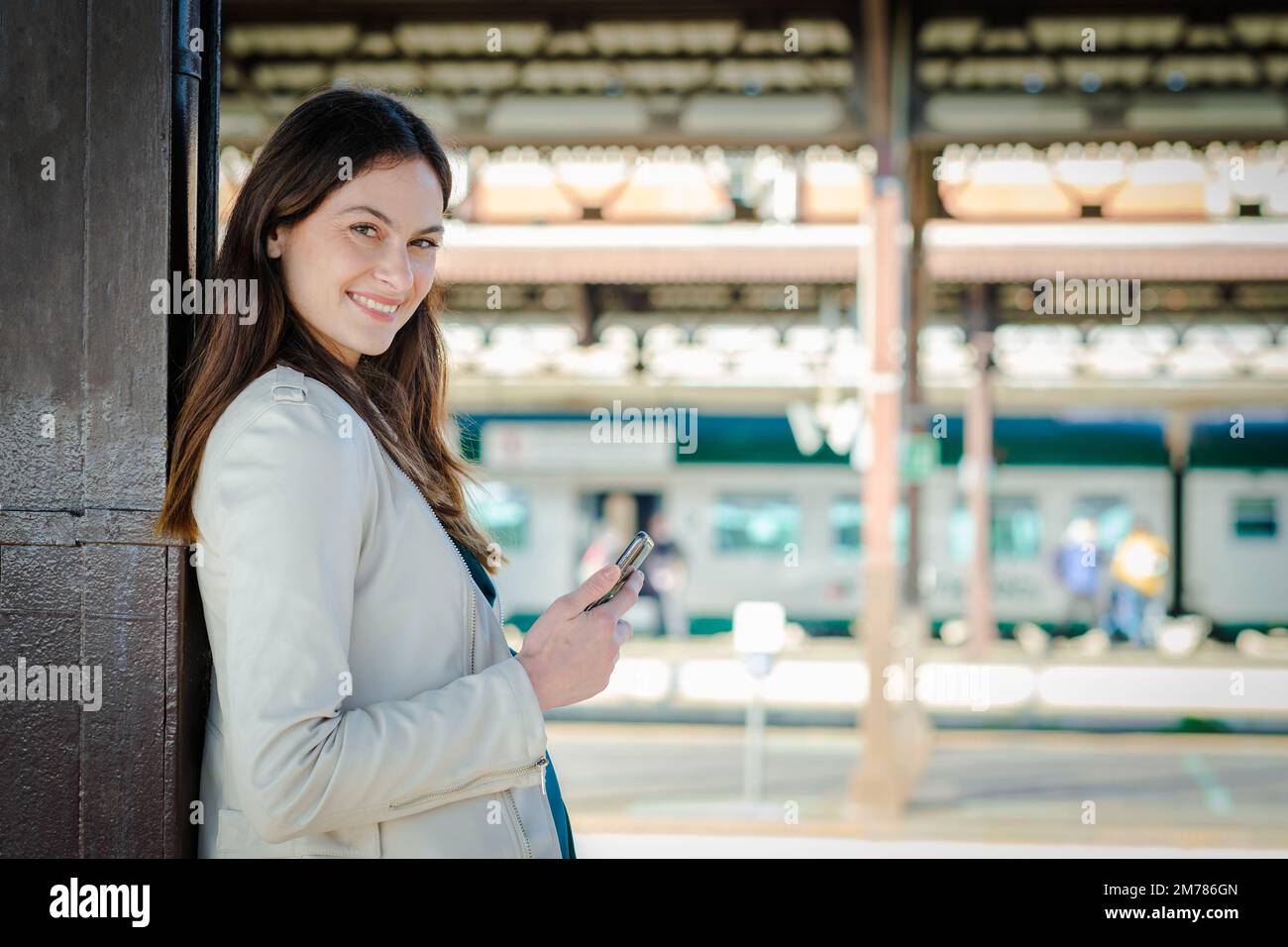 Femme d'affaires souriante utilisant un smartphone en attendant dans une gare - jeune fille étudiante attrayante utilisant le téléphone portable en position debout Banque D'Images