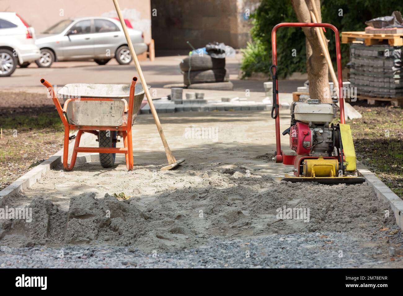 Une brouette de construction et un compacteur de sol à essence sur un fond flou d'un chantier de pose de revêtement par une journée d'été. Banque D'Images