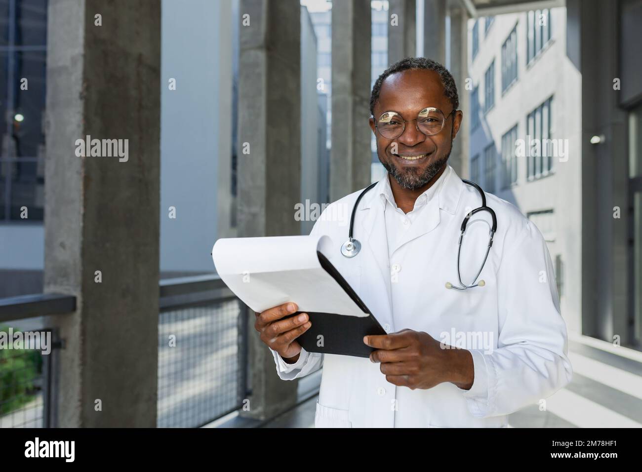Portrait d'un beau médecin afro-américain. Il est debout près de l'hôpital sous un manteau blanc avec une chemise dans ses mains, regardant la caméra, souriant. Banque D'Images