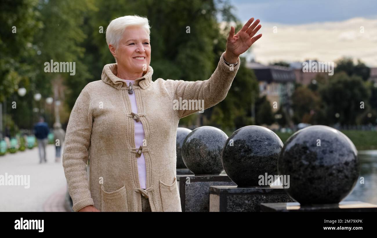 Portrait d'une femme d'âge souriant et belle qui regarde loin en ordonnant bonjour ou Au revoir se détendant à l'extérieur par temps frais, heureuse femme sénior aux cheveux gris Banque D'Images