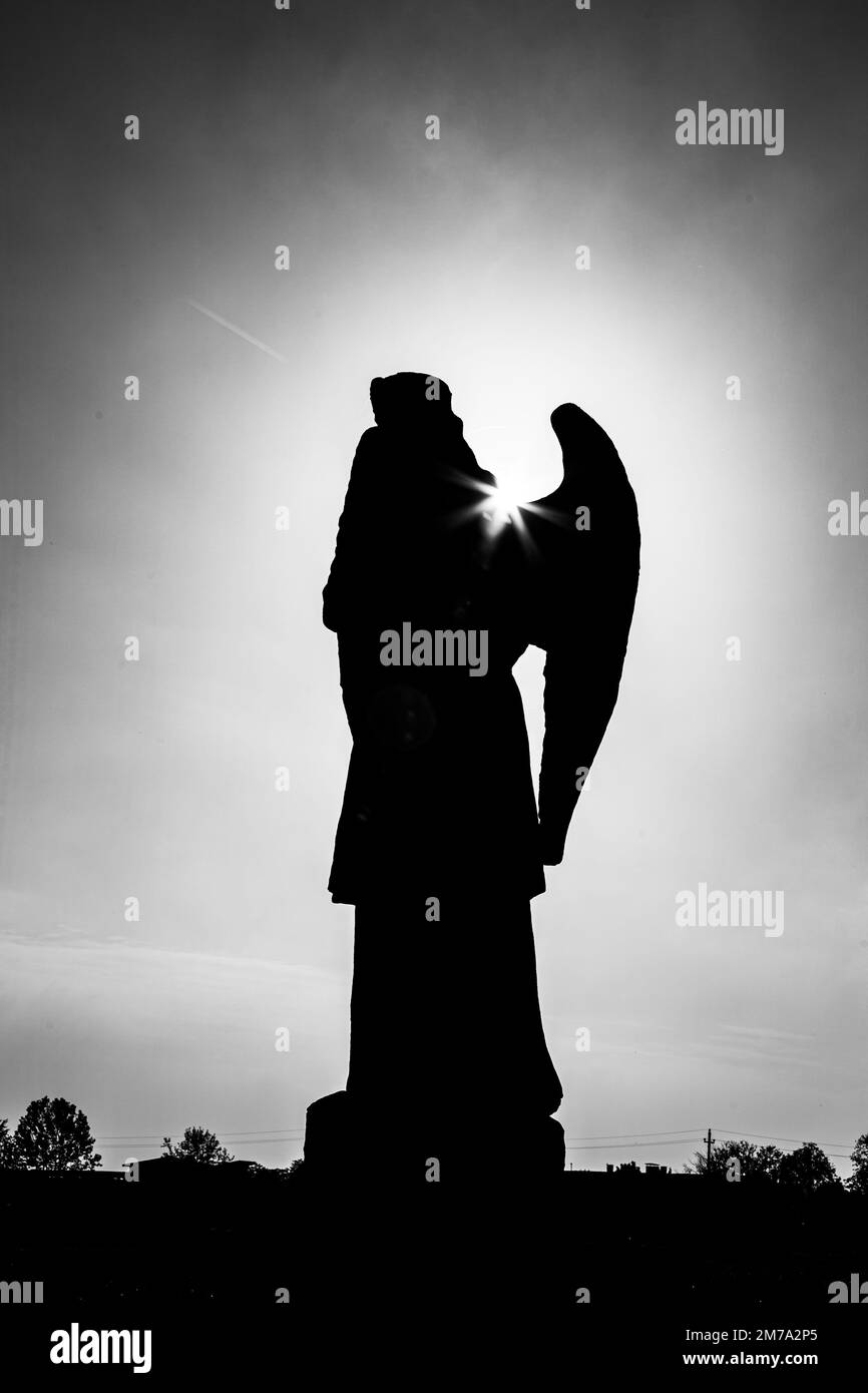 Une photo en niveaux de gris de la silhouette d'une statue d'ange avec des ailes sous ciel nuageux au coucher du soleil Banque D'Images
