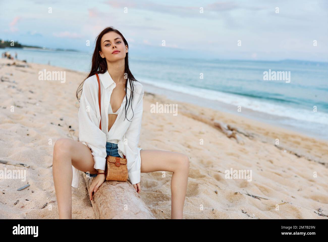 Femme de mode assise sur le sable dans une chemise blanche et short près de l'océan avec un sac posant sur les vacances, la mode et la beauté, peau de bronzage Banque D'Images