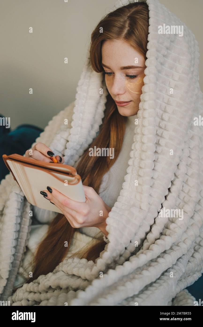 Une belle jeune femme enveloppée dans une couverture assise sur un canapé pensant et écrivant des notes. Portrait contemporain. Appartement confortable. Banque D'Images