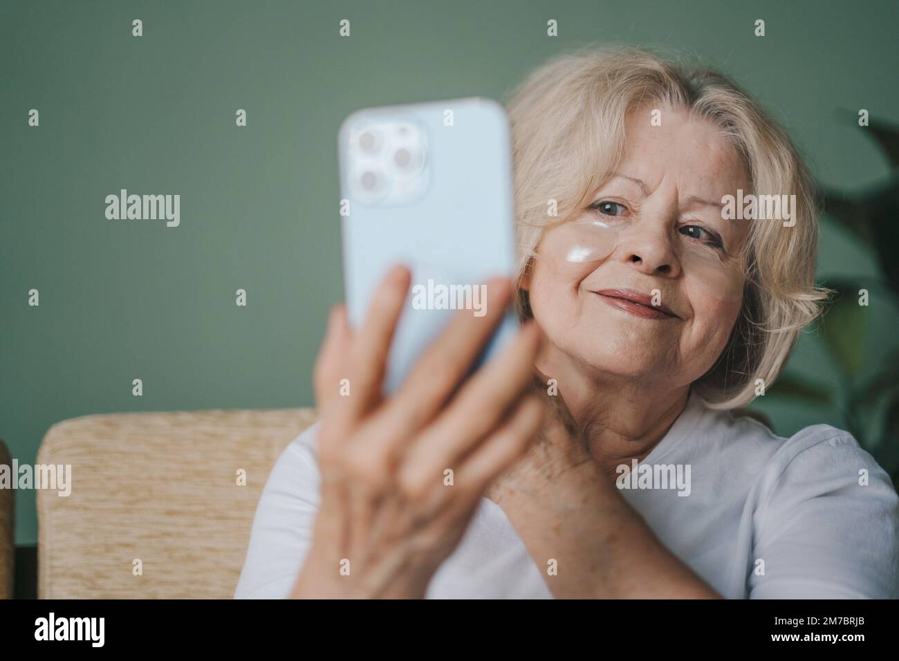 Femme sénior attirante tenant le smartphone devant elle, en utilisant l'écran comme miroir tout en appliquant des cellules oculaires sous ses yeux. Smartphone. Internet Banque D'Images