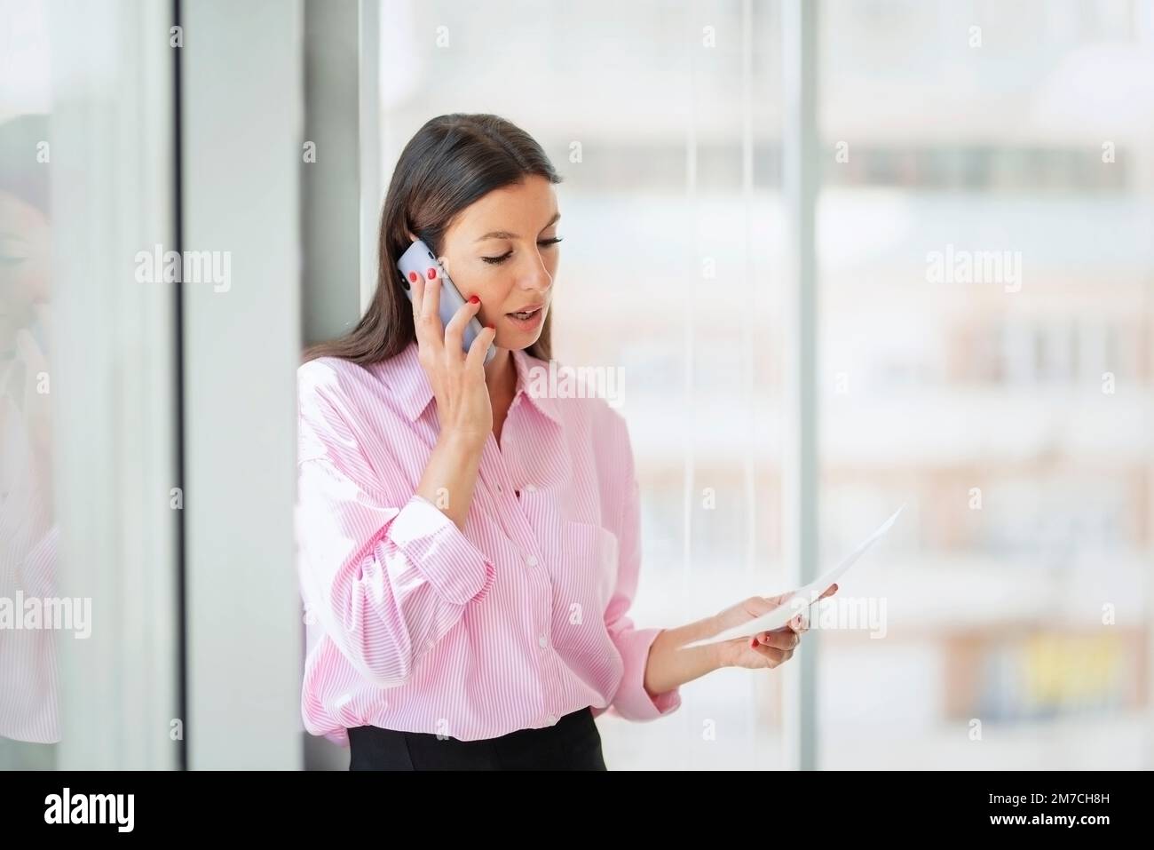 Femme d'affaires souriante debout au bureau tout en utilisant un smartphone et en tenant un document dans la main. Femme professionnelle ayant un appel d'affaires. Banque D'Images