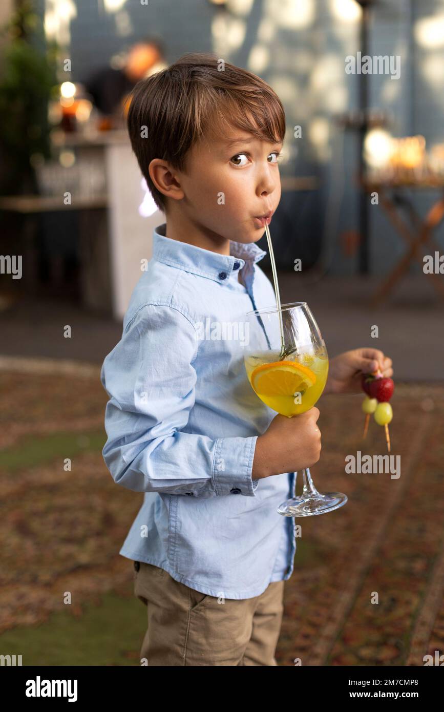 Portrait d'un petit garçon buvant du jus dans un verre, décoré de fruits, avec de la paille dans le parc extérieur. Enfant en été Banque D'Images