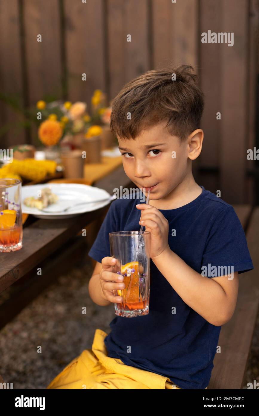 Portrait d'un petit garçon buvant du jus dans un verre, décoré de fruits, avec de la paille dans le jardin extérieur. Enfant en été Banque D'Images