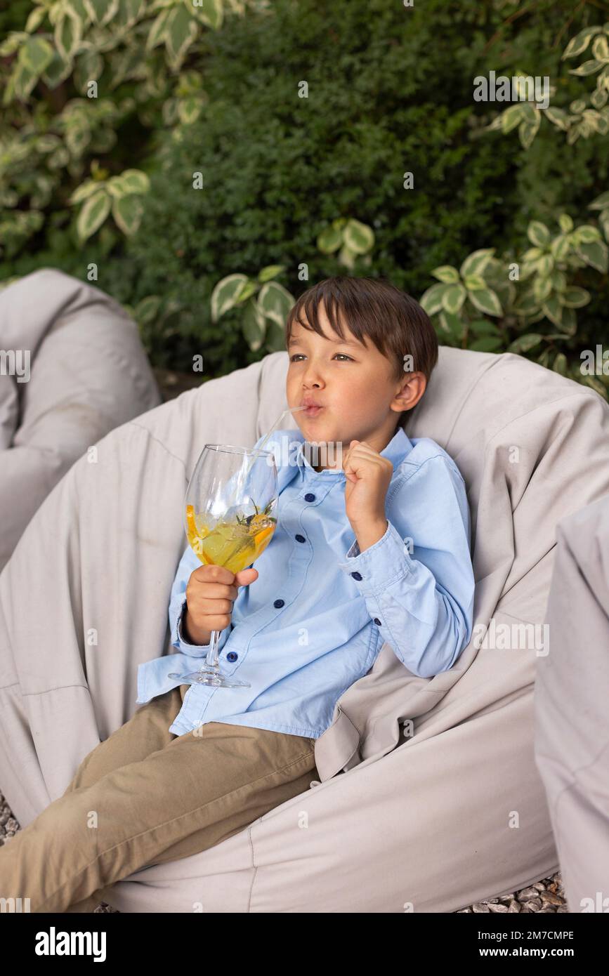 Portrait d'un petit garçon buvant du jus dans un verre, décoré de fruits, avec de la paille dans le parc extérieur. Enfant en été Banque D'Images