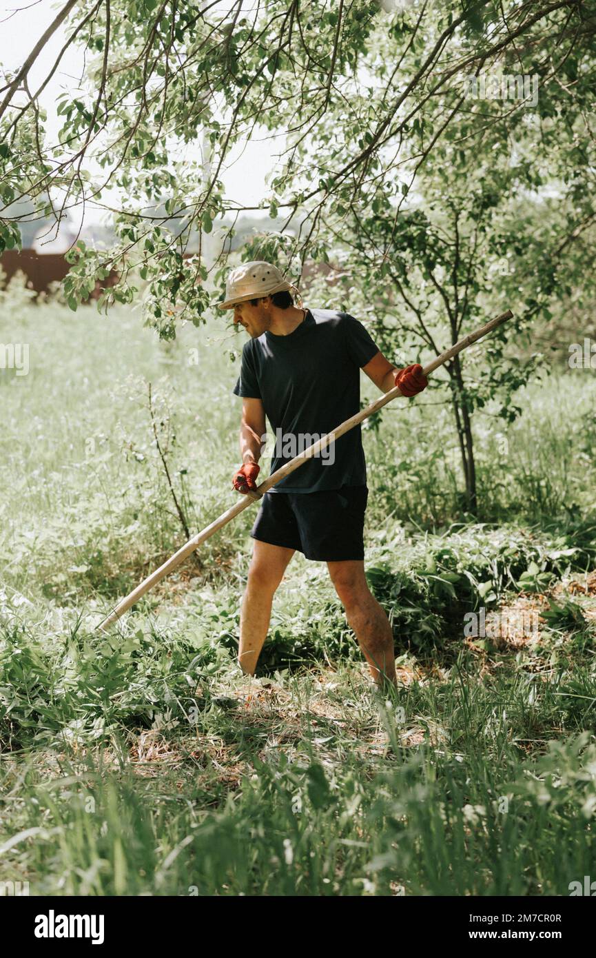 faucher l'herbe traditionnelle à l'ancienne avec la main scythe sur la ferme du village domestique. jeune agriculteur adulte fauchant le vert de l'herbe cultivée Banque D'Images