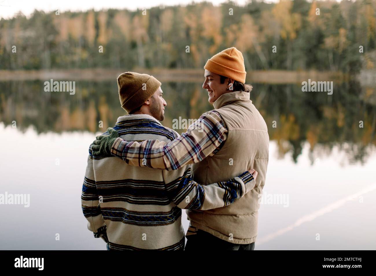 Des amis masculins parlent les uns avec les autres tout en se tenant avec le bras autour contre le lac Banque D'Images
